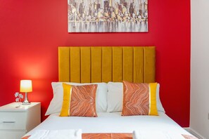 A hotel room with a red wall, a cityscape painting, a bed with white and orange pillows, and a nightstand with a lamp. - Modern 5 Bed Flat in Thornaby Road For Big Groups (Stockton-on-Tees)