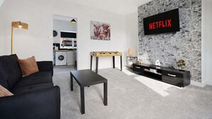 Una sala de estar moderna con un sofá negro, una mesa de centro, mesa de fútbol, televisor montado en una pared de piedra y una cocina al fondo.