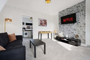 A modern living room with a black sofa, a coffee table, a foosball table, a TV mounted on a stone wall, and a kitchen in the background. - Horden 2BR Home Sleeps 6 By Stay With us (Peterlee)