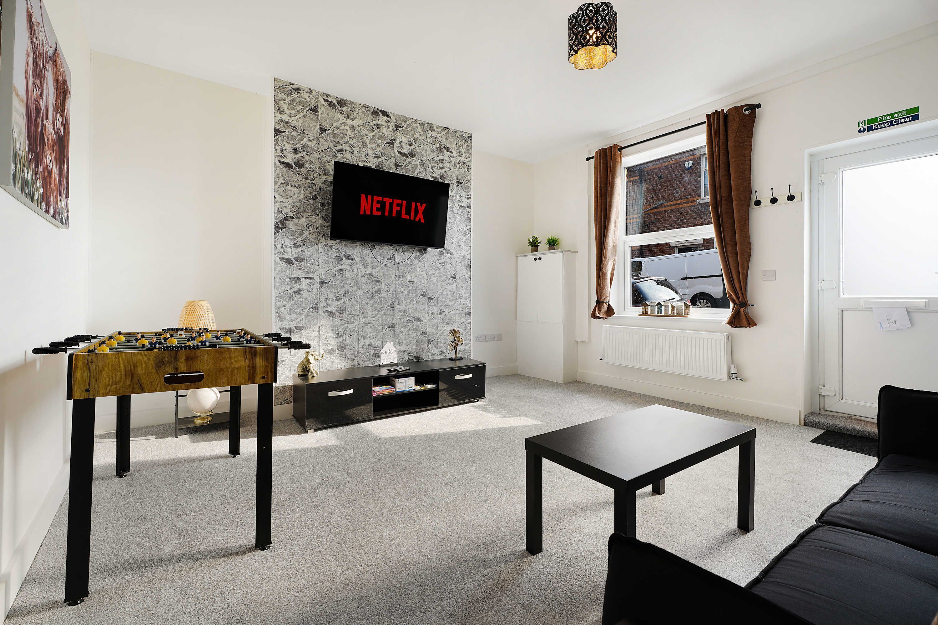 A living room with a foosball table, a flat-screen TV displaying 'NETFLIX', a sofa, a coffee table, and a window with curtains.
