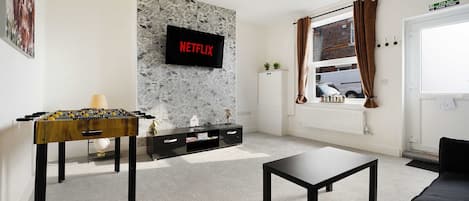 A living room with a foosball table, a flat-screen TV displaying 'NETFLIX', a sofa, a coffee table, and a window with curtains.