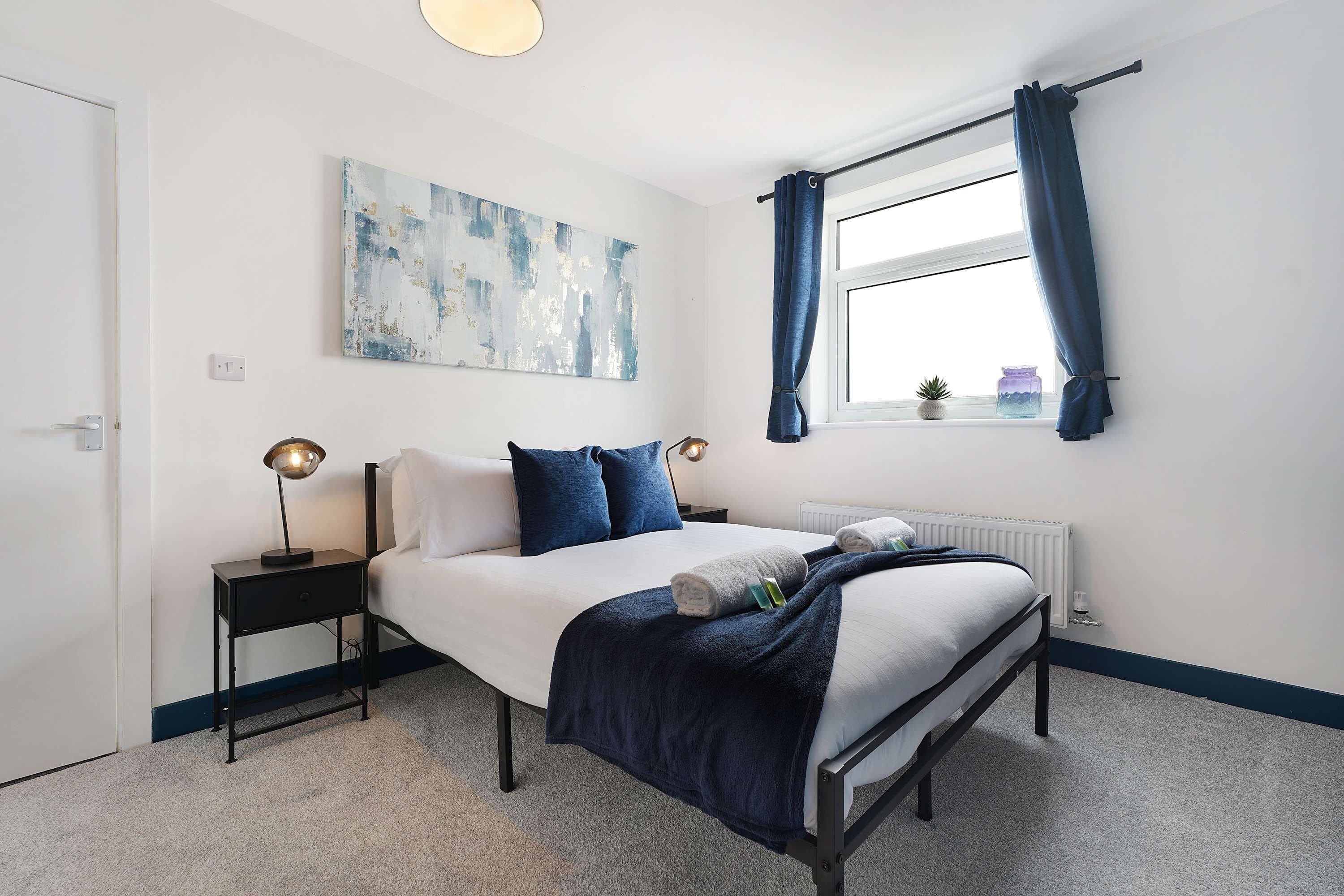 A bedroom with a bed, a nightstand, a lamp, a window with curtains, a radiator, and a wall-mounted artwork.