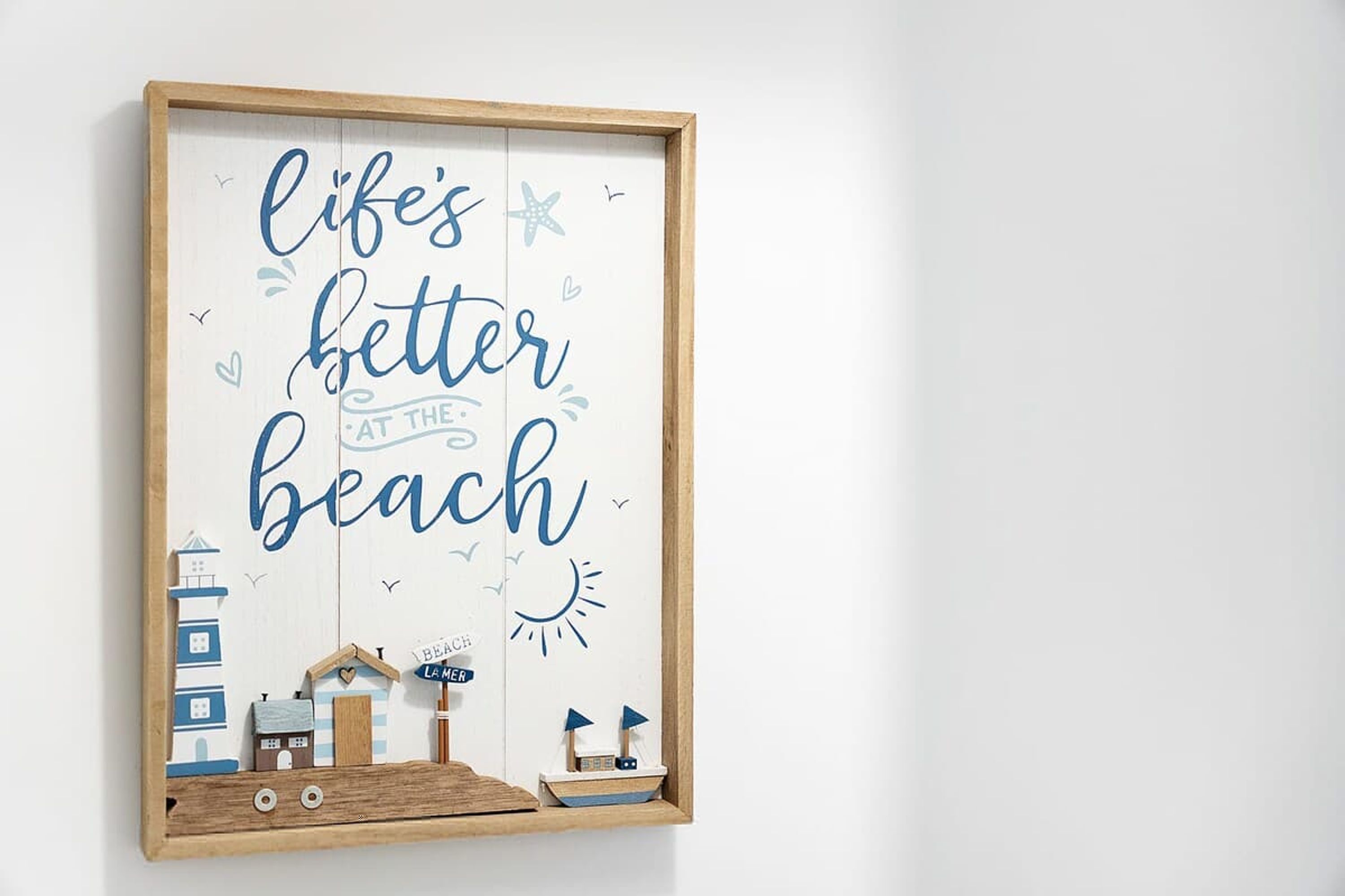 A framed sign with the text 'life's better at the beach' and decorative elements like a lighthouse, a house, and a boat.