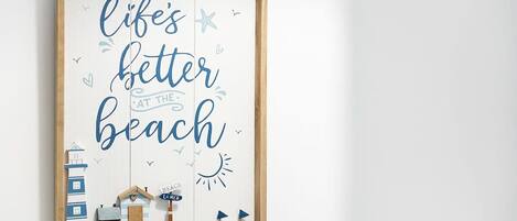 A framed sign with the text 'life's better at the beach' and decorative elements like a lighthouse, a house, and a boat.