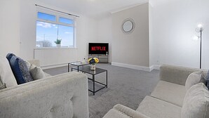 A modern living room with a sofa, a coffee table, a TV displaying 'NETFLIX', and a large window. - Newly Refurbished Near the Coast Sleeps 5 (Newbiggin-by-the-Sea)