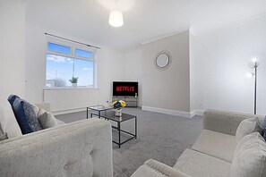 Ein modernes Wohnzimmer mit einer Couch, einem Couchtisch, einem Fernseher mit 'NETFLIX' und einem großen Fenster.