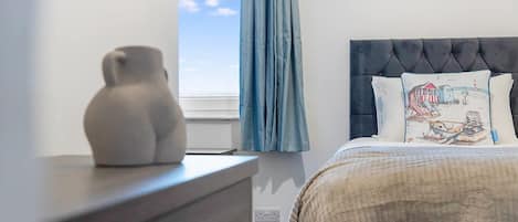A bedroom with a bed, a dresser, a window with curtains, and a view of the sky.