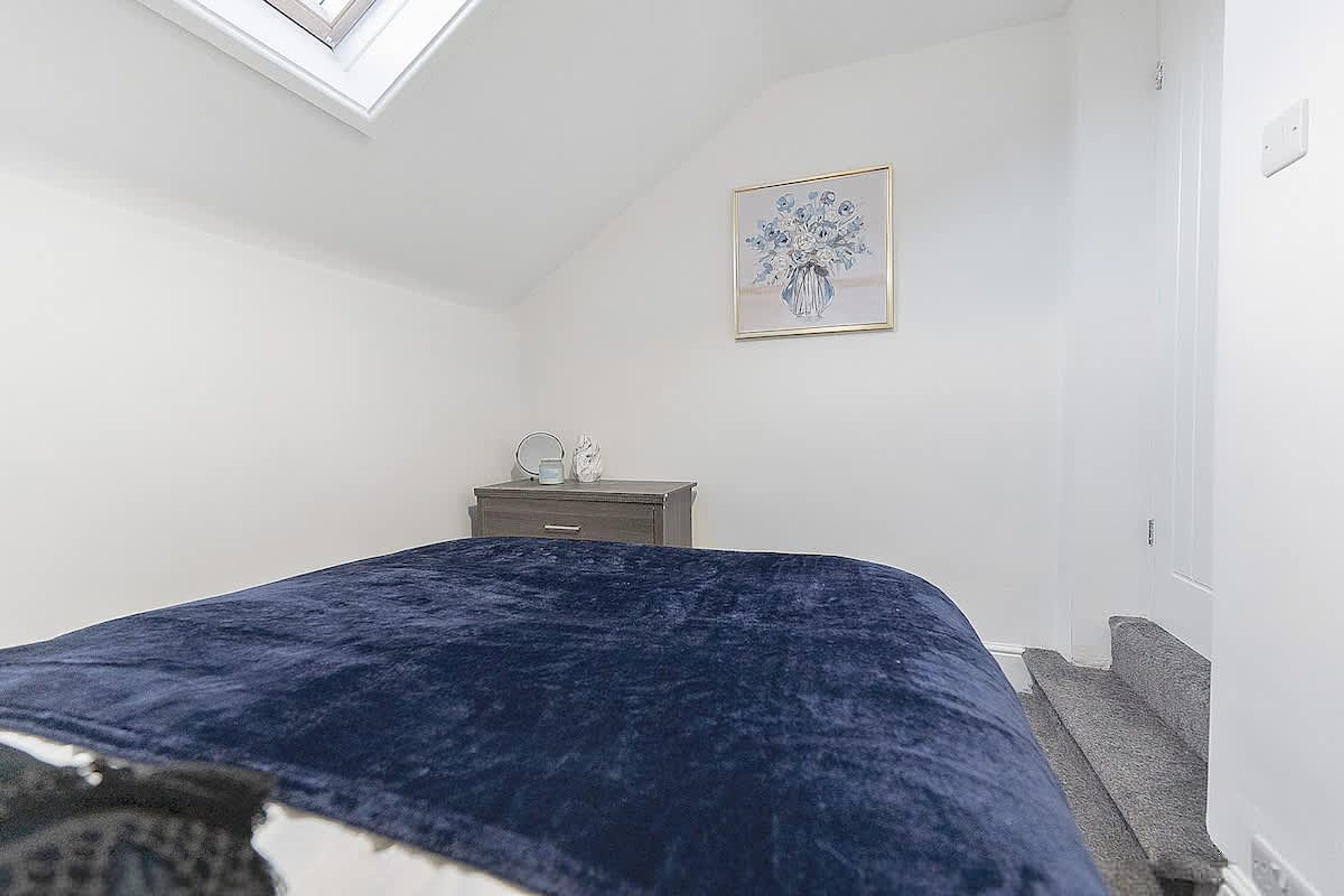 Une chambre à coucher avec un lit, une commode, une table de chevet, une fenêtre et un tableau accroché au mur.