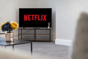 Ein Wohnzimmer mit einem Fernseher, der 'NETFLIX' anzeigt, einem Couchtisch mit einem Blumenstrauß und einem Teppichboden.