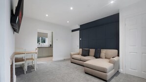 Ein modernes Wohnzimmer mit einem beigefarbenen Sofa, einem Esstisch mit Stühlen, einer Küchenzeile und einem flachbildfernseher an der Wand.