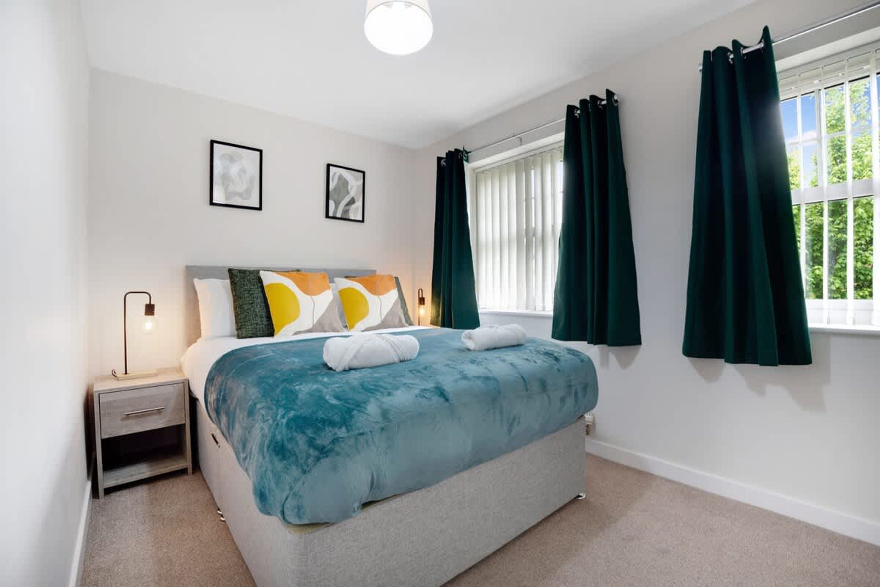 A bedroom with a bed, a nightstand, a window with curtains, and two framed pictures on the wall.