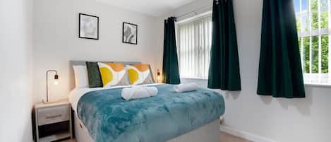 A bedroom with a bed, a nightstand, a window with curtains, and two framed pictures on the wall.