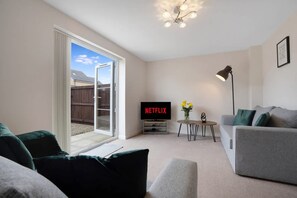 A living room with a grey sofa, a TV displaying 'NETFLIX', a small table with a vase, and a ceiling light fixture. - Modern Stockton Retreat Sleeps 6 by Stay With Us (Stockton-on-Tees)