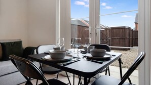 A dining area with a table set for four, chairs, and a view of a backyard with a wooden fence and a building. - Modern Stockton Retreat Sleeps 6 by Stay With Us (Stockton-on-Tees)