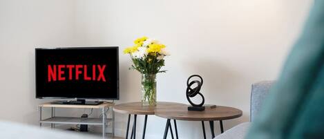 Una sala de estar con un televisor mostrando el logo de Netflix, un florero con flores y una escultura moderna sobre una mesita.