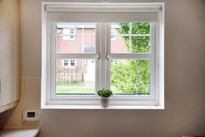 A window with a white frame, a small potted plant on the sill, and a view of a brick building and greenery outside. - Modern Stockton Retreat Sleeps 6 by Stay With Us (Stockton-on-Tees)