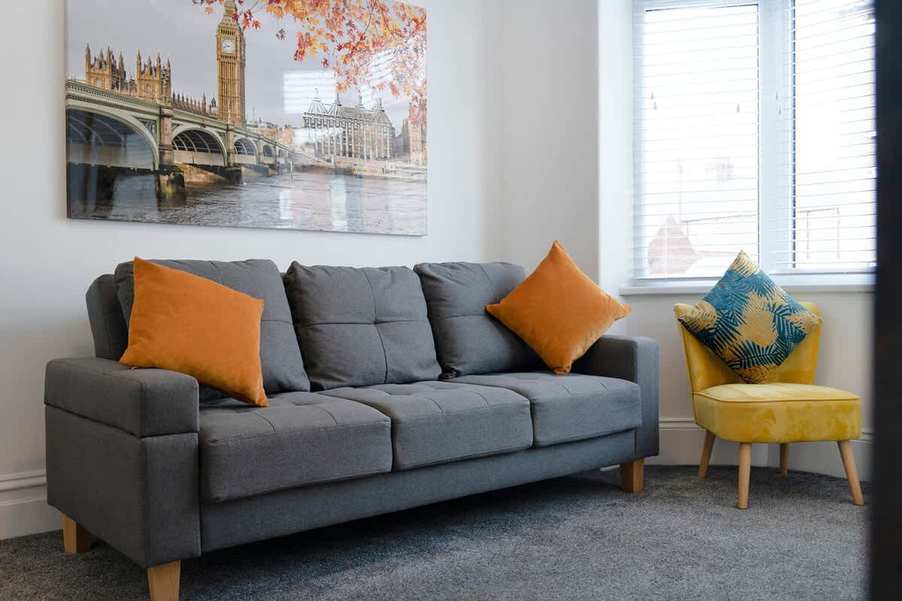 A grey sofa with two orange cushions and a yellow chair with a patterned cushion in a room with a large window and a cityscape painting on the wall.