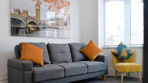A grey sofa with two orange cushions and a yellow chair with a patterned cushion in a room with a large window and a cityscape painting on the wall. - 2BR Darlington Home by Stay With Us (Darlington)