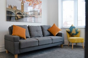 A grey sofa with two orange cushions and a yellow chair with a patterned cushion in a room with a large window and a cityscape painting on the wall. - 2BR Darlington Home by Stay With Us (Darlington)