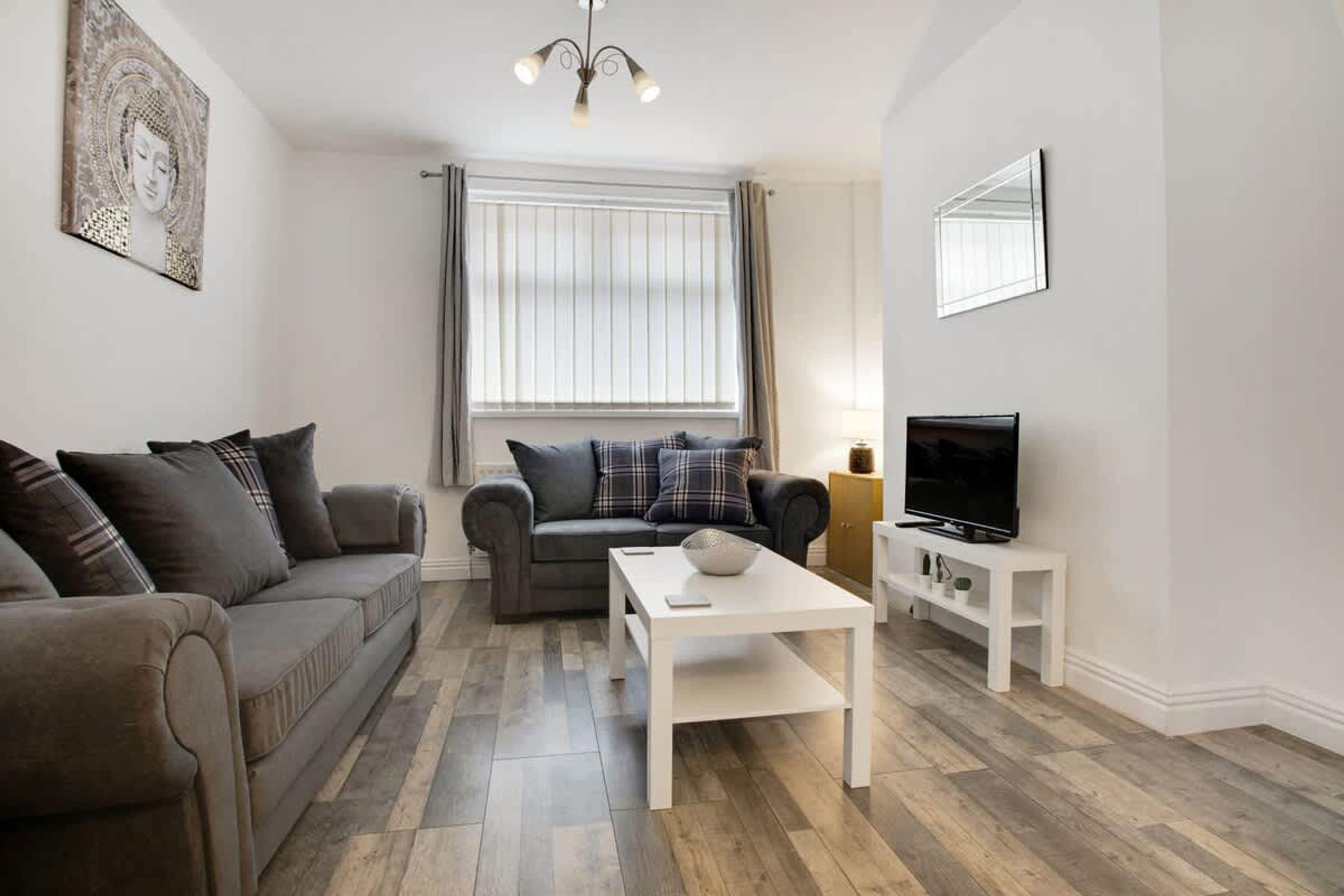 A living room with a grey sofa, a white coffee table, a television on a stand, and a window with curtains.