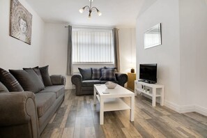 Un salon avec un canapé gris, une table basse blanche, un téléviseur posé sur un meuble et une fenêtre avec des rideaux.