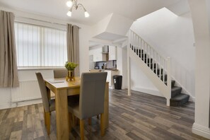 Un coin repas avec une table et des chaises en bois, une cuisine à l’arrière-plan et un escalier menant à l’étage supérieur.