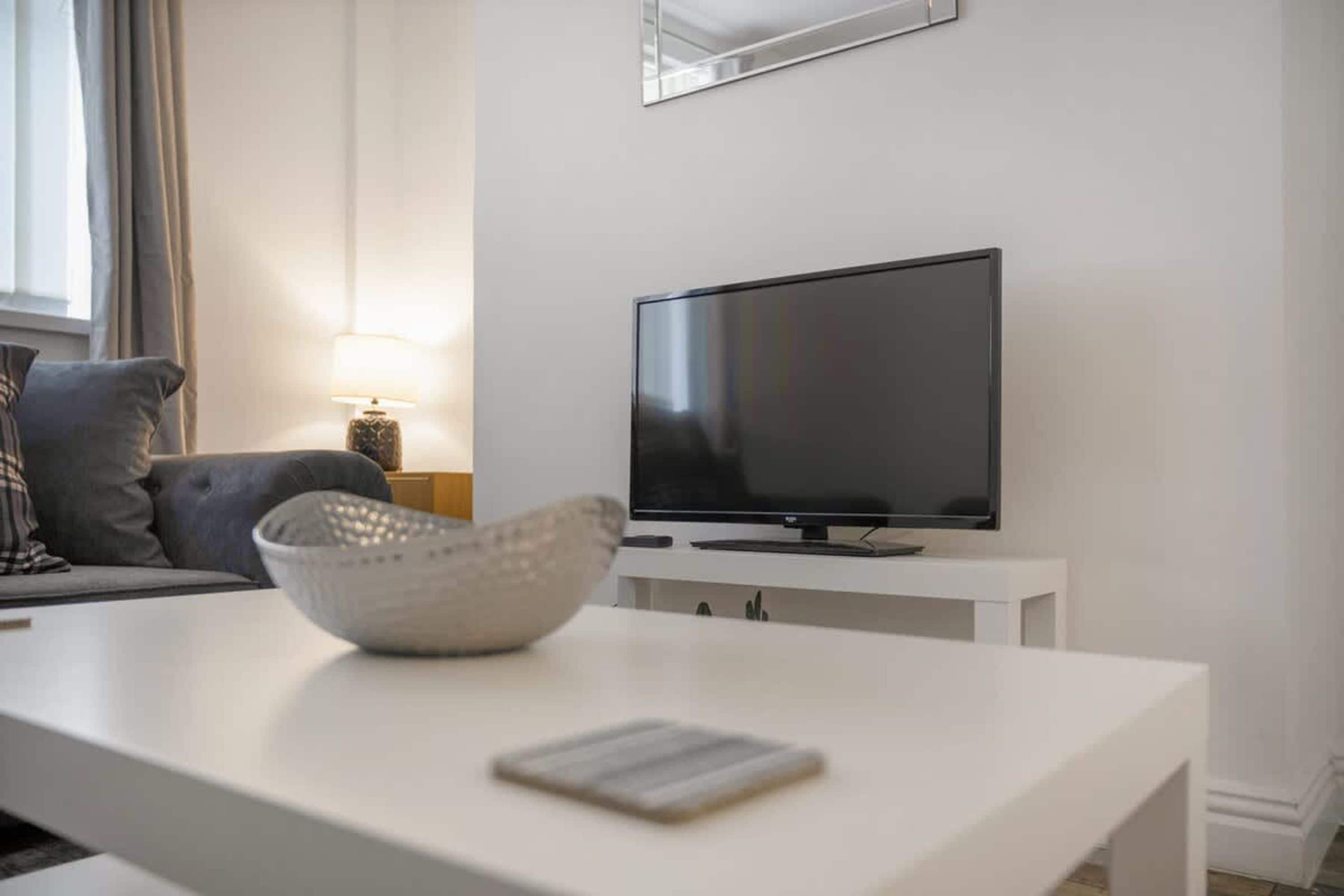 A modern living room with a flat-screen TV on a white stand, a grey sofa, a lamp, and a white coffee table with a bowl and a book.