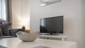 Una sala de estar moderna con un televisor de pantalla plana sobre un soporte blanco, un sofá gris, una lámpara y una mesa de centro blanca con un cuenco y un libro.