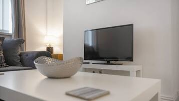 A modern living room with a flat-screen TV on a white stand, a grey sofa, a lamp, and a white coffee table with a bowl and a book.