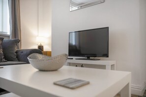 Ein modernes Wohnzimmer mit einem Flachbildfernseher auf einem weißen Standfuß, einem grauen Sofa, einer Lampe und einem weißen Couchtisch mit einer Schale und einem Buch.