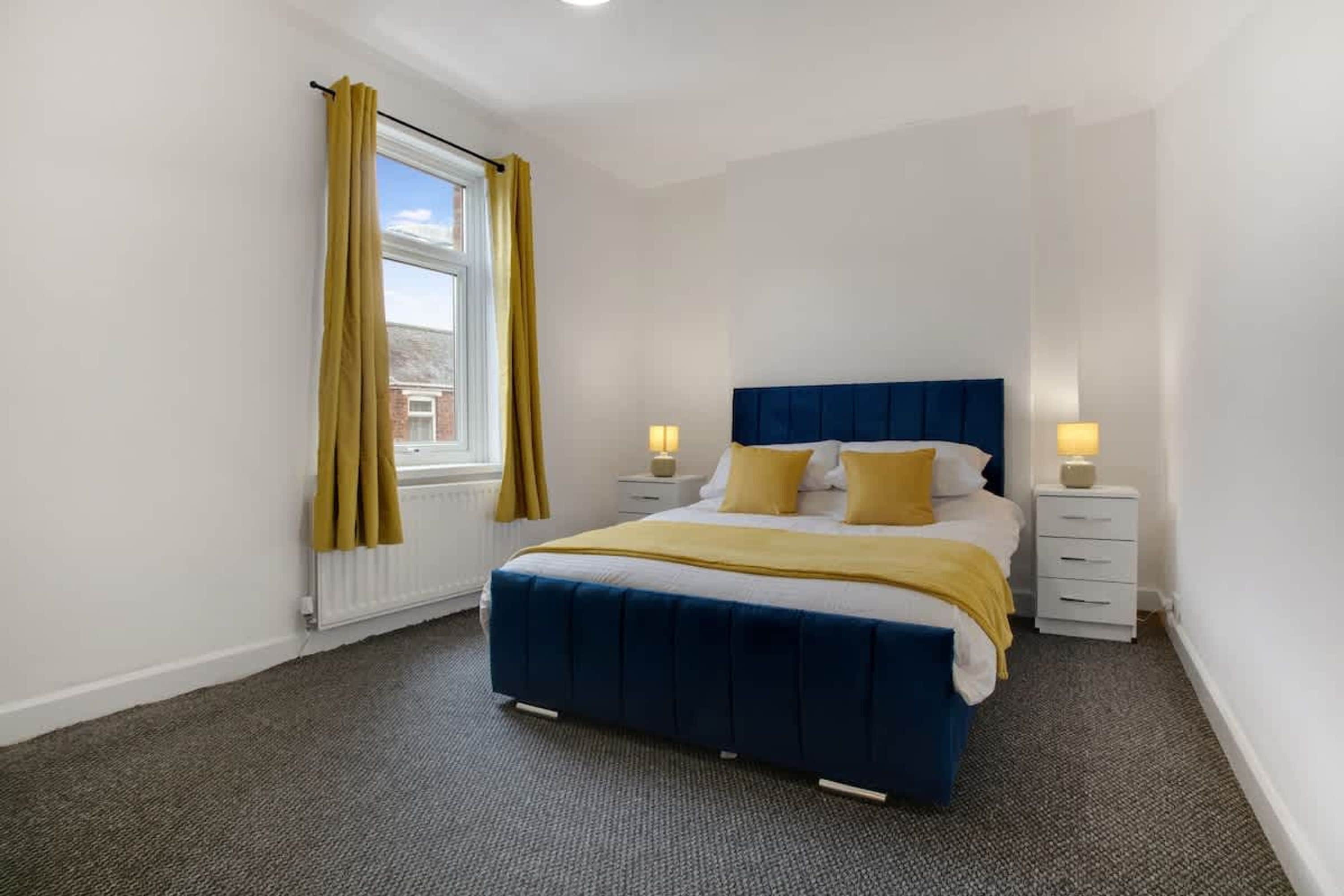 A bedroom with a bed, two bedside tables, a window with curtains, and a radiator.