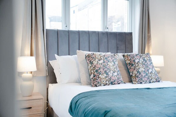 Una cama bien hecha con una colcha azul y dos almohadas florales, flanqueada por dos lámparas de noche sobre mesitas de madera.