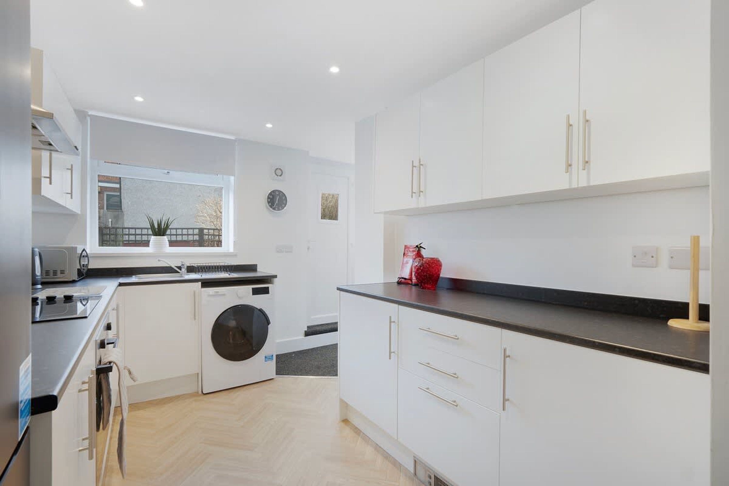 A modern kitchen with white cabinetry, a built-in washing machine, and a window overlooking a balcony.