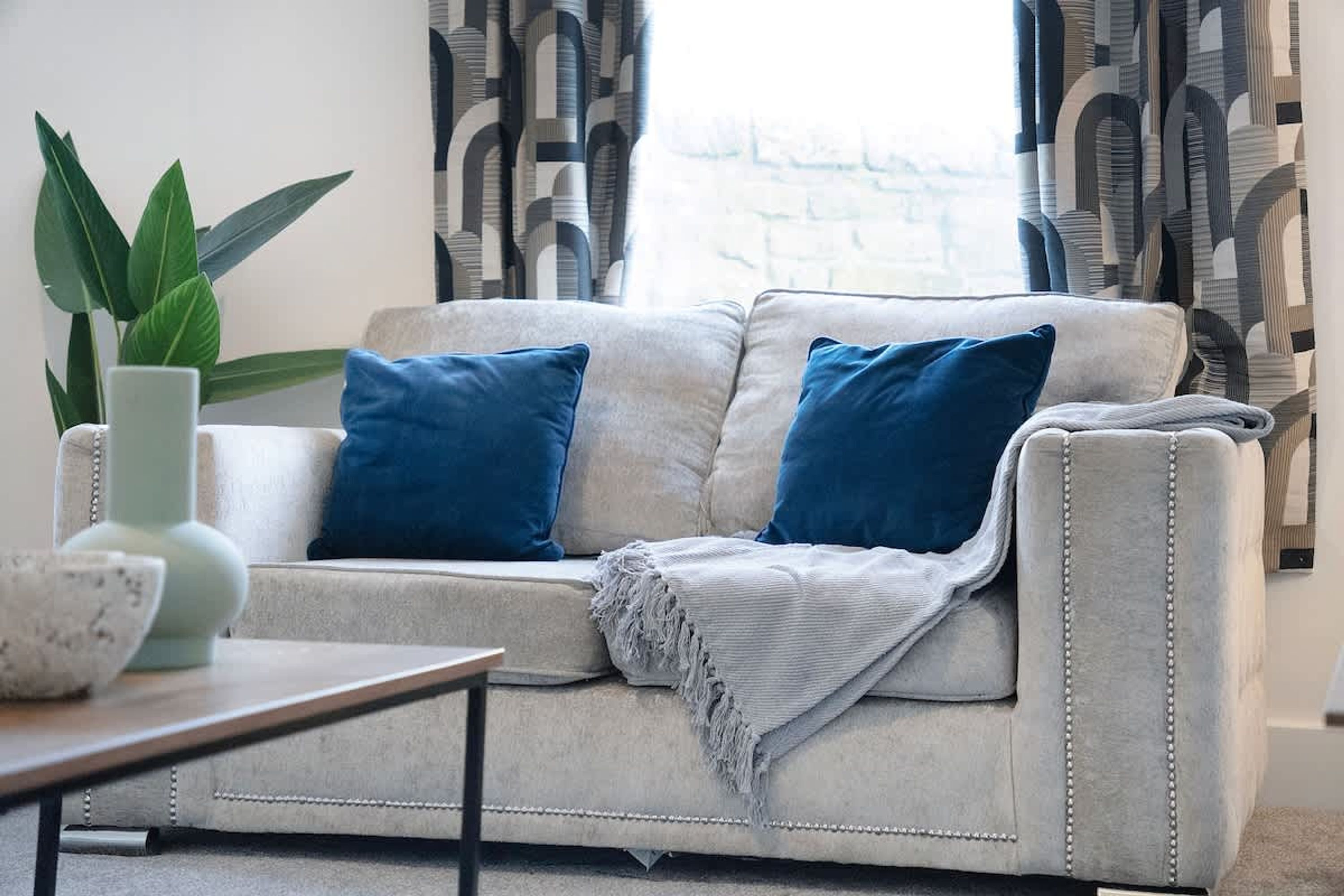 A living room with a sofa, two blue cushions, a green plant, and a patterned curtain.