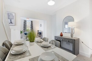 Un comedor con una mesa lista para cuatro personas, un sofá, un espejo y un aparador con una lámpara.