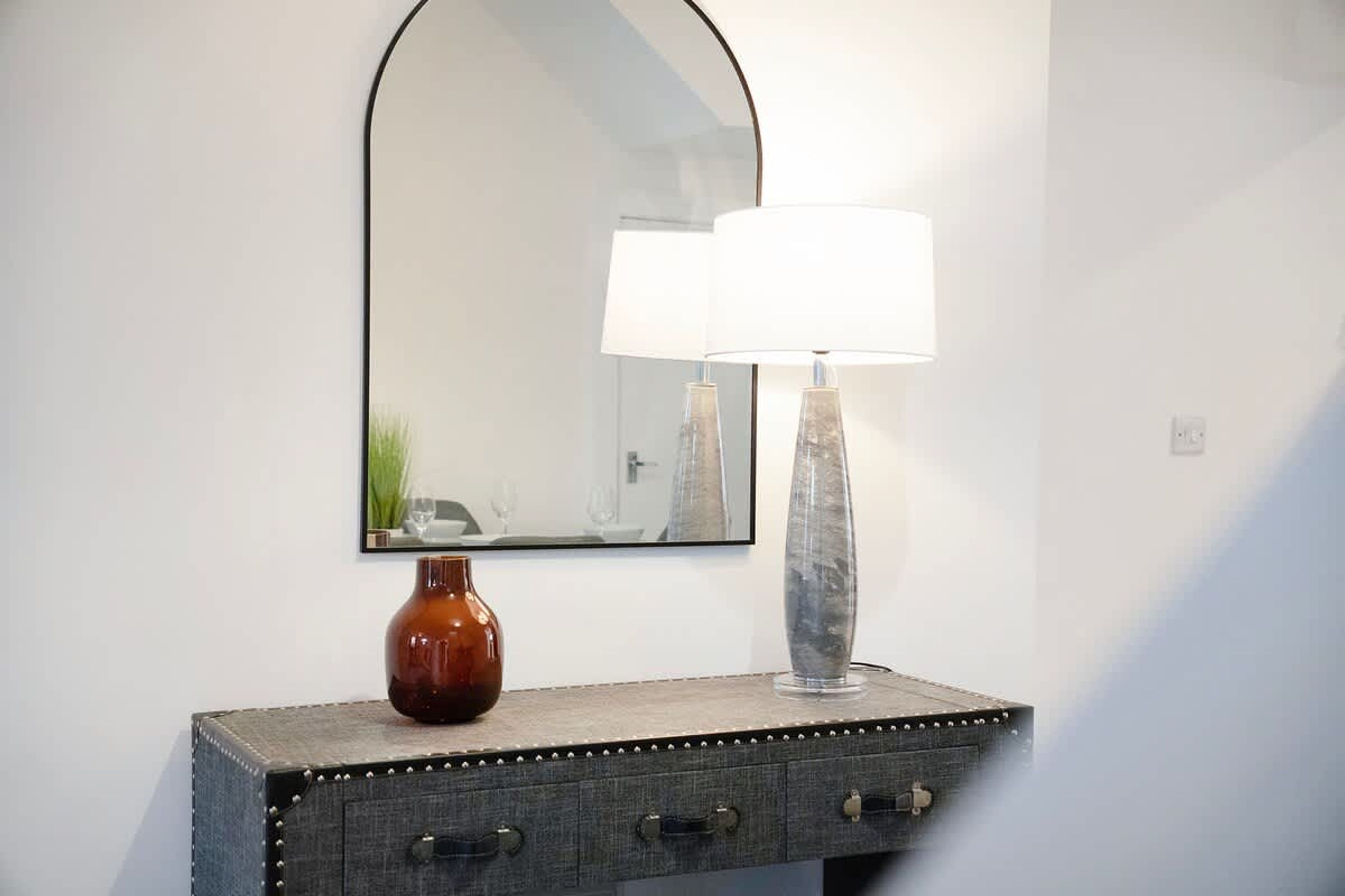 A mirror with a simple black frame, a vase with a white lampshade, and a small, dark-colored cabinet with two drawers.