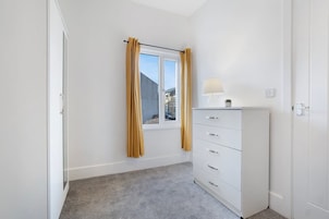 Una habitación con ventana, un aparador blanco con cajones, una lámpara encima y una alfombra gris.