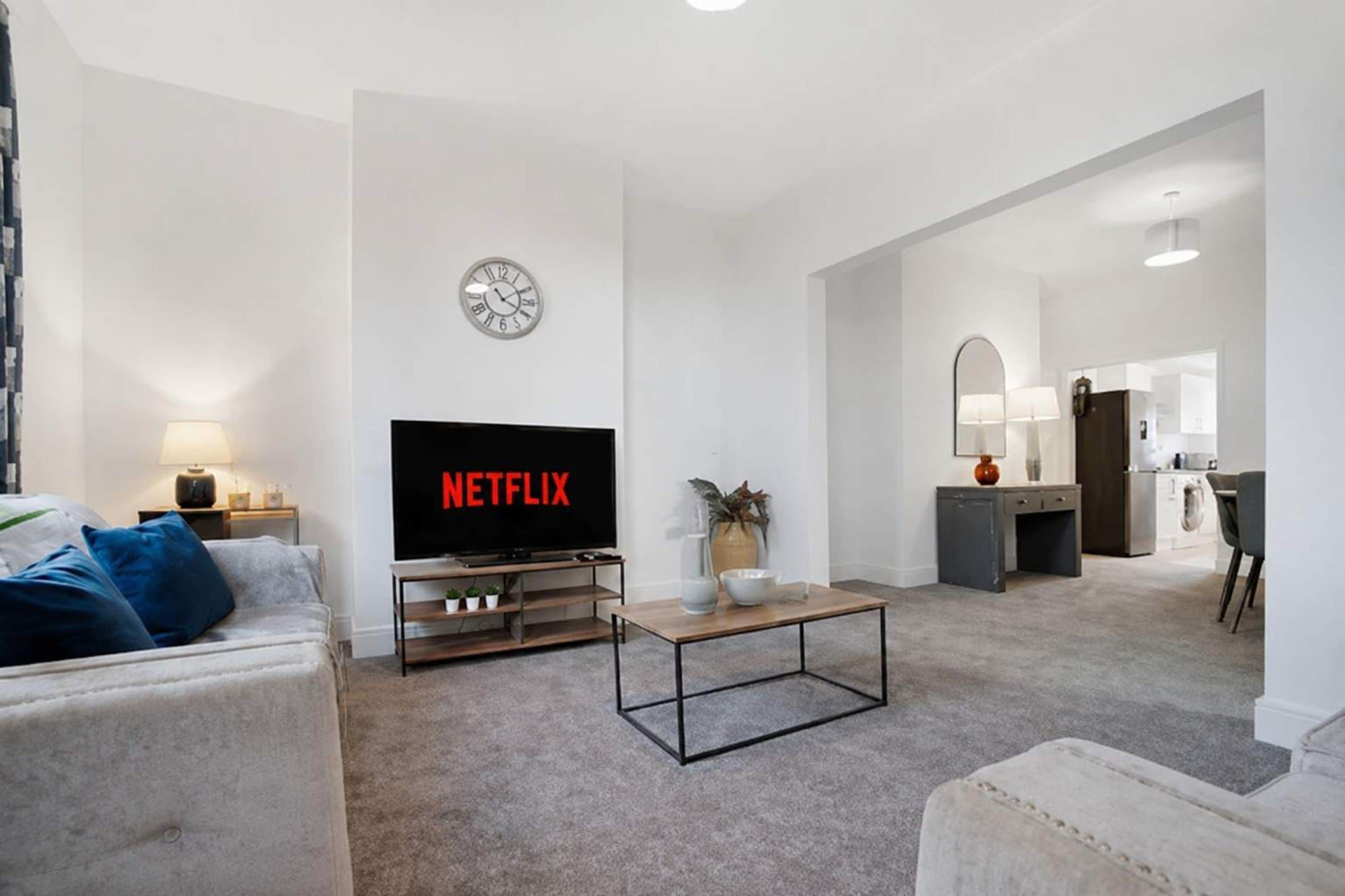 A modern living room with a flat-screen TV displaying 'NETFLIX,' a clock on the wall, and a kitchen area in the background.