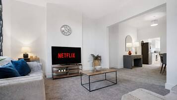 A modern living room with a flat-screen TV displaying 'NETFLIX,' a clock on the wall, and a kitchen area in the background.