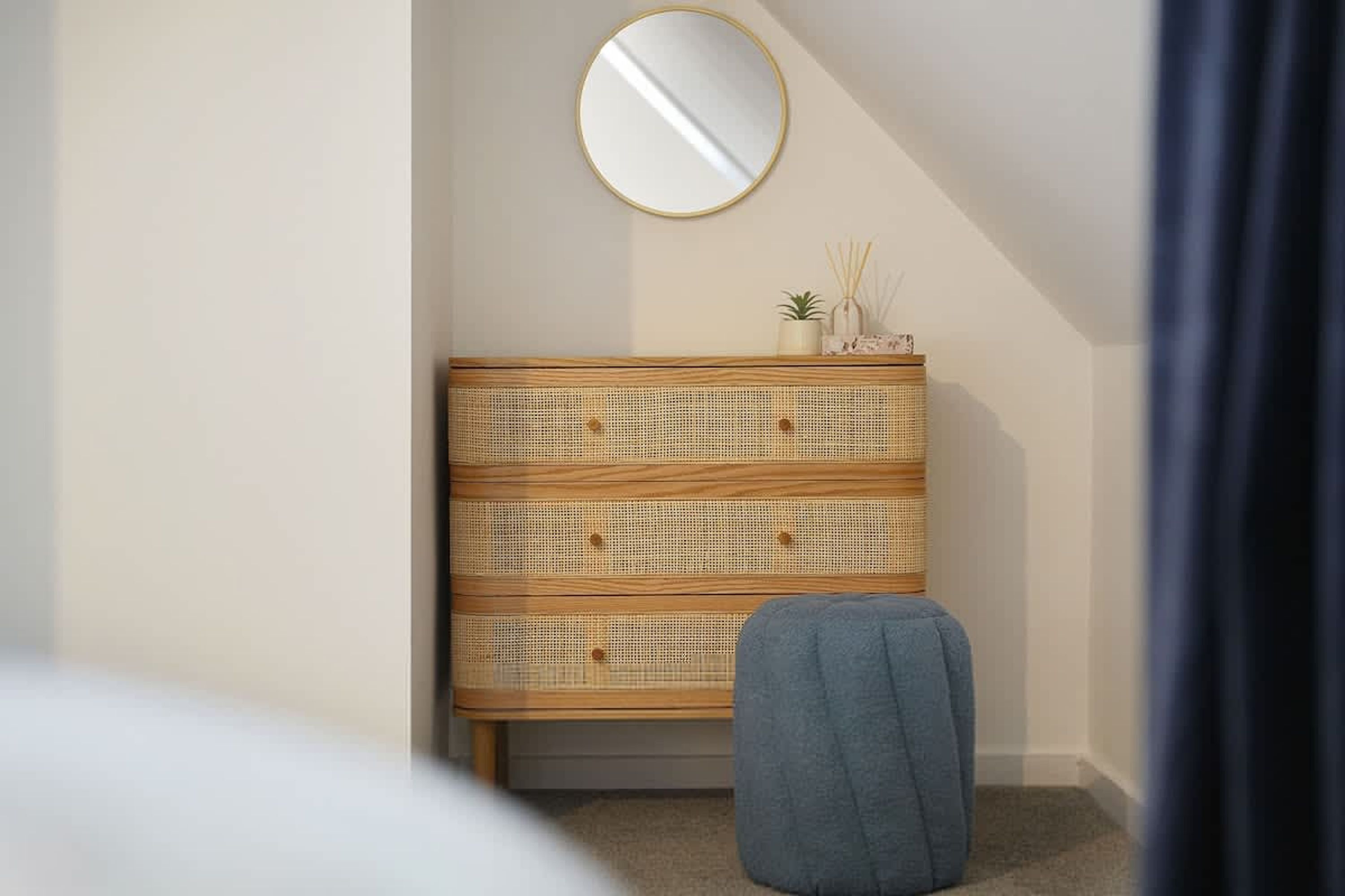Une pièce avec une commode en bois, un miroir rond et un pouf bleu.