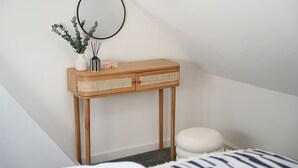 Habitación de esquina con una mesita consola de madera, un espejo redondo, un taburete blanco pequeño y una cama con manta a rayas.
