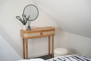 Ein Eckschlafzimmer mit einem Holzkonsolentisch, einem runden Spiegel, einem kleinen weißen Hocker und einem Bett mit gestreifter Tagesdecke.