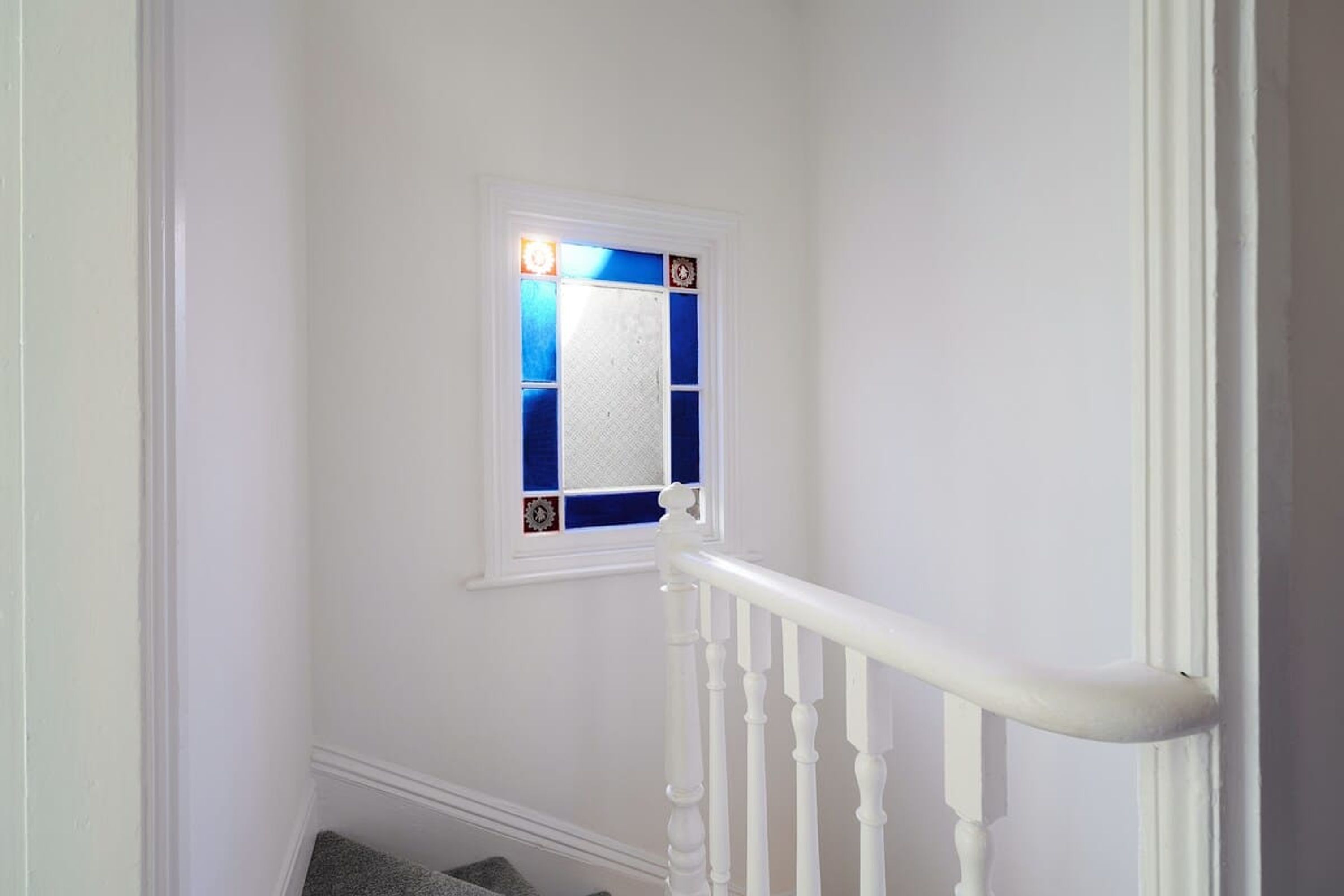 Un escalier blanc avec une rampe blanche et une petite fenêtre aux carreaux bleus, agrémentée d’éléments décoratifs.