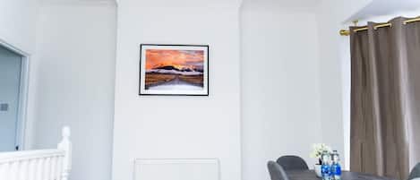 Ein Zimmer mit weißer Wand, einem gerahmten Bild, einem Heizkörper, einem Esstisch mit Stühlen und einem Fenster mit Vorhängen.