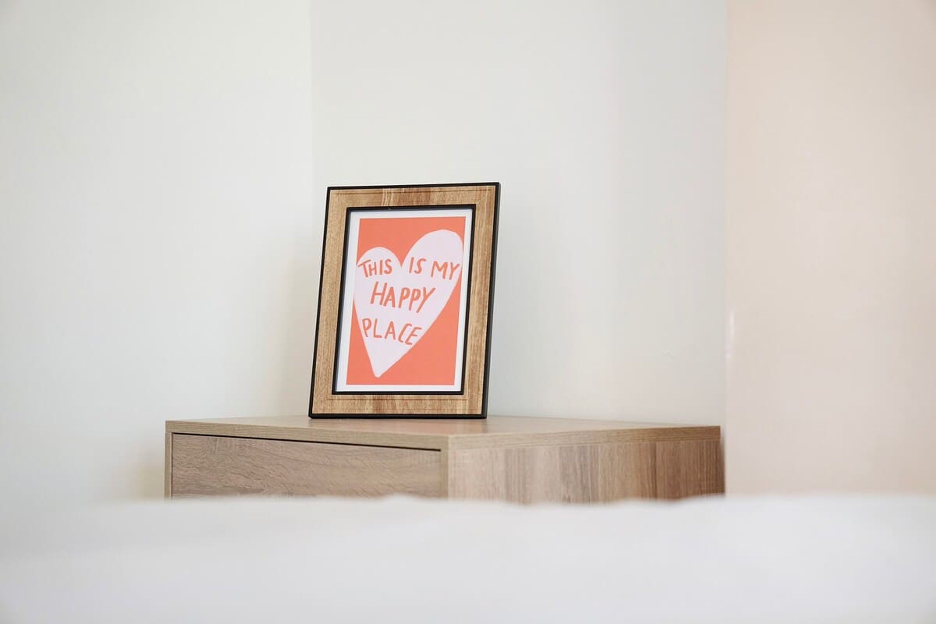 A framed sign with the text 'THIS IS MY HAPPY PLACE' on a wooden surface against a plain wall.