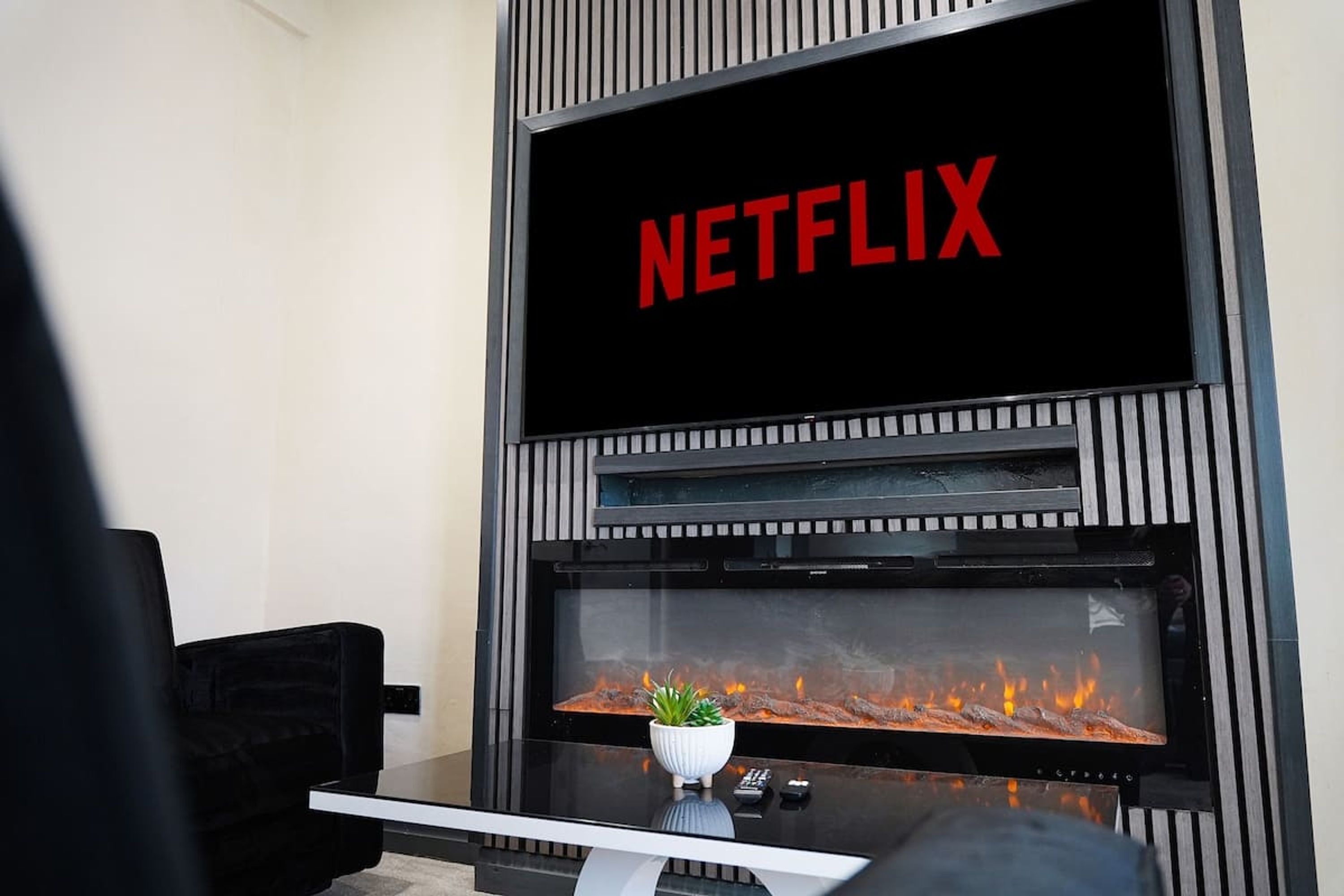 A modern living room with a fireplace, a television mounted above it displaying the Netflix logo, and a small plant on the mantel.