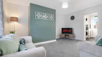Moderni olohuone, jossa on harmaa matto, valkoiset sohvat, litteänäyttöinen televisio, jossa näkyy teksti ”NETFLIX”, ja seinään kiinnitetty valaisin.