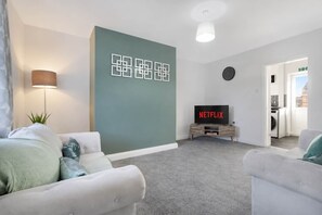 A modern living room with a grey carpet, white sofas, a flat-screen TV displaying 'NETFLIX', and a wall-mounted lamp. - Relaxing 2BR in Billy Row Wifi by Stay With Us (Crook)