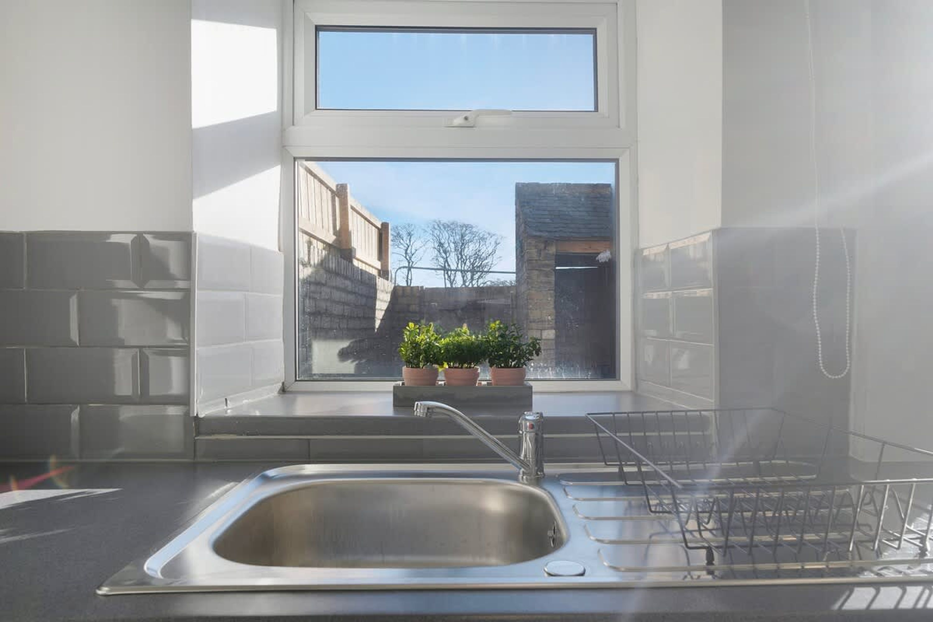 Uma cozinha com uma pia de aço inoxidável, uma janela com vista para edifícios e plantas em vasos no parapeito.