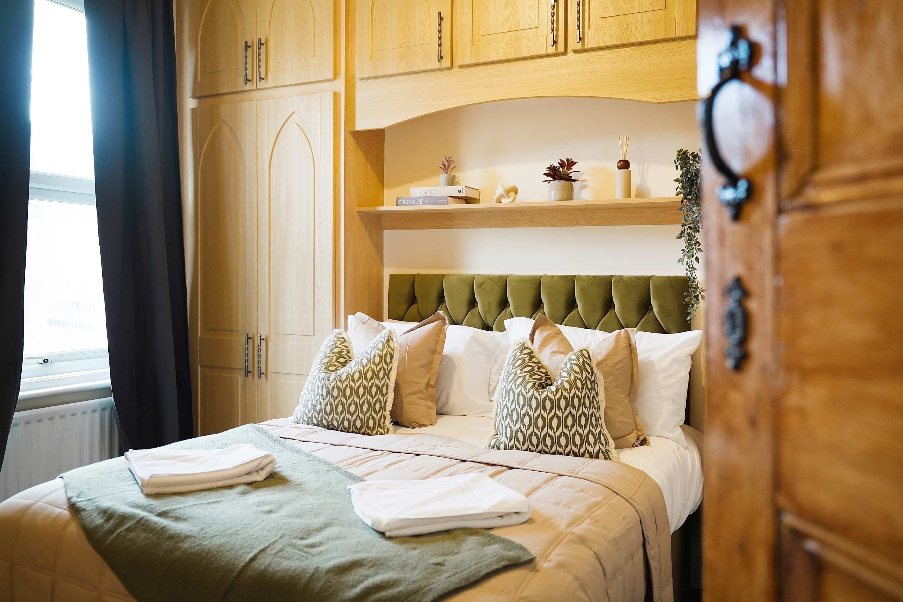 A hotel room with a bed, a window with curtains, wooden cabinets, and a shelf with decorative items.